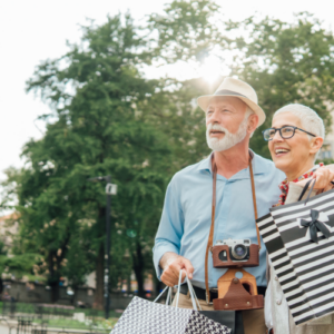 An elderly couple with several shopping bags strolling in a park while the man has a digital camera hanging from his neck.