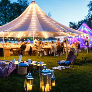 An evening event with beautiful white tents and lighting with a lot of people gathered at the long table underneath the tent. 