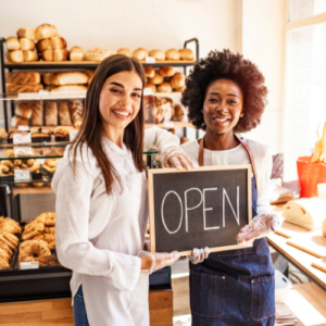 Two women holding an "Open" sign in a bread shop