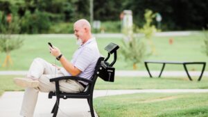 Senior male, sitting on a solar charging bench, charging his phone while actively using it.